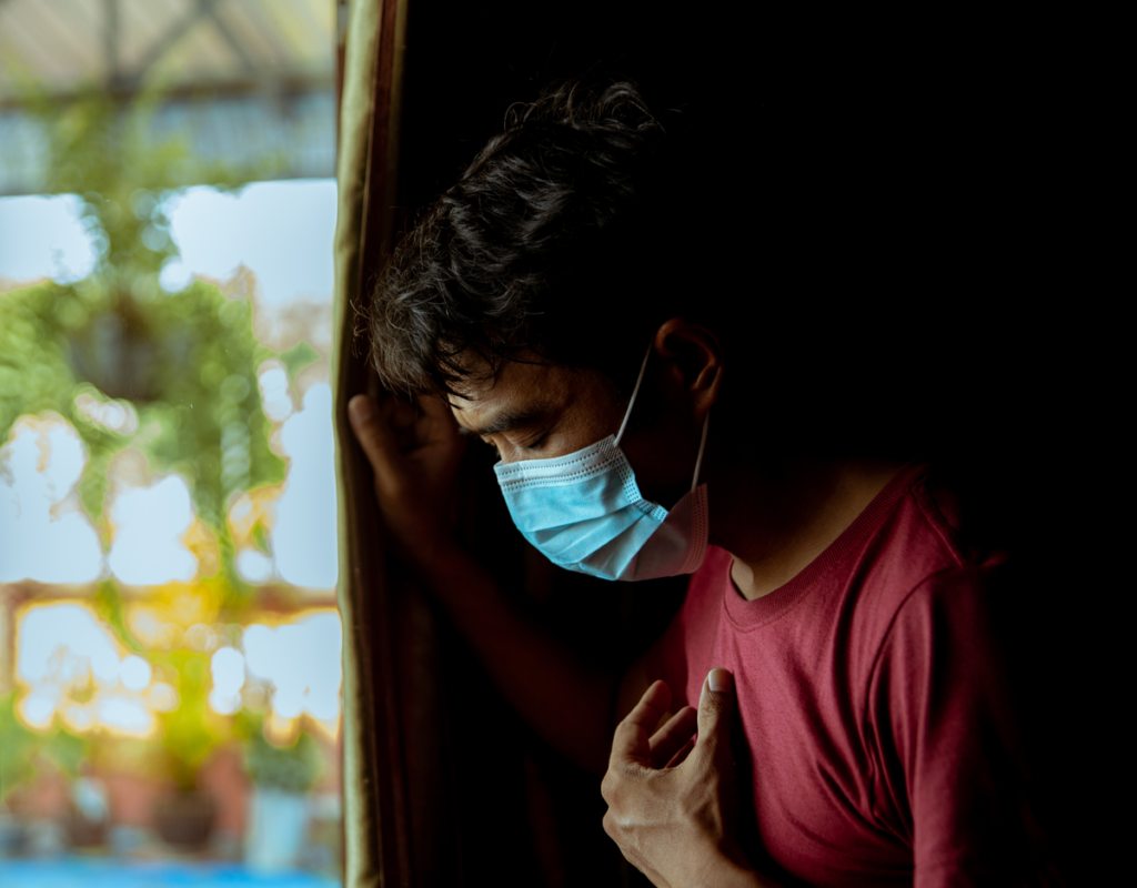 A man stands at a window with a blue mask on. He is holding his chest as he breathes. He has COVID.